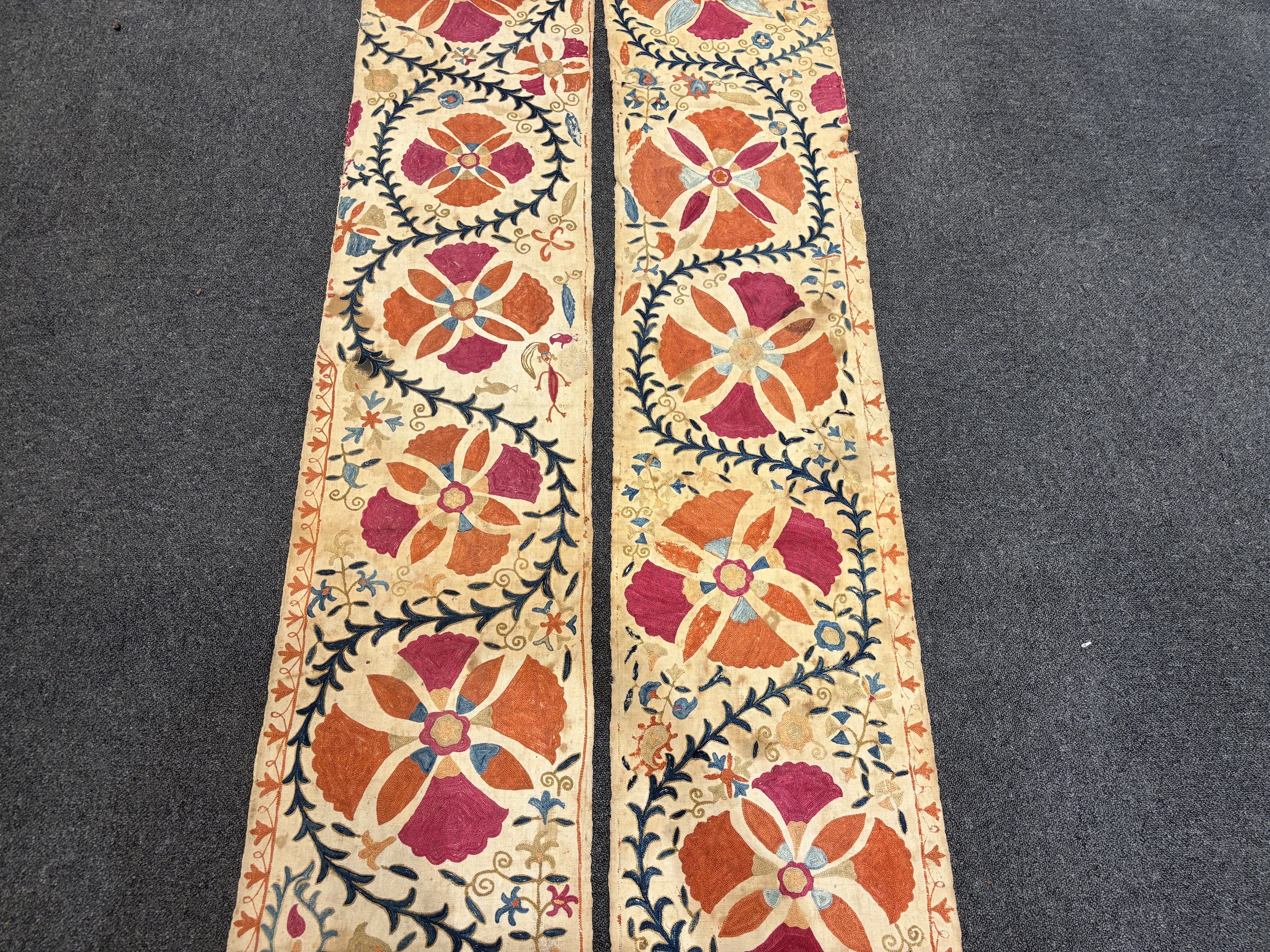 A pair of Uzbekistan long wide panels, both embroidered in mostly red, coral and blue with large symbolic flowers and trailing vines on woven cotton using mostly Burkara couching, longest 250cm long x 38cm wide